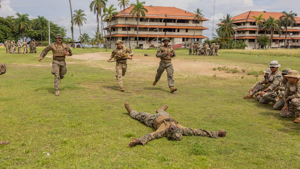 Military personnel are conducting a training exercise on a grass field near a multi-story building with a red-tiled roof, surrounded by palm trees in what appears to be a tropical location. One soldier is lying prone on the ground while three others run toward him, suggesting a casualty response or combat drill scenario. A group of additional soldiers observes from the right side, some crouching and recording with a phone.
