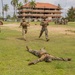 Military personnel are conducting a training exercise on a grass field near a multi-story building with a red-tiled roof, surrounded by palm trees in what appears to be a tropical location. One soldier is lying prone on the ground while three others run toward him, suggesting a casualty response or combat drill scenario. A group of additional soldiers observes from the right side, some crouching and recording with a phone.