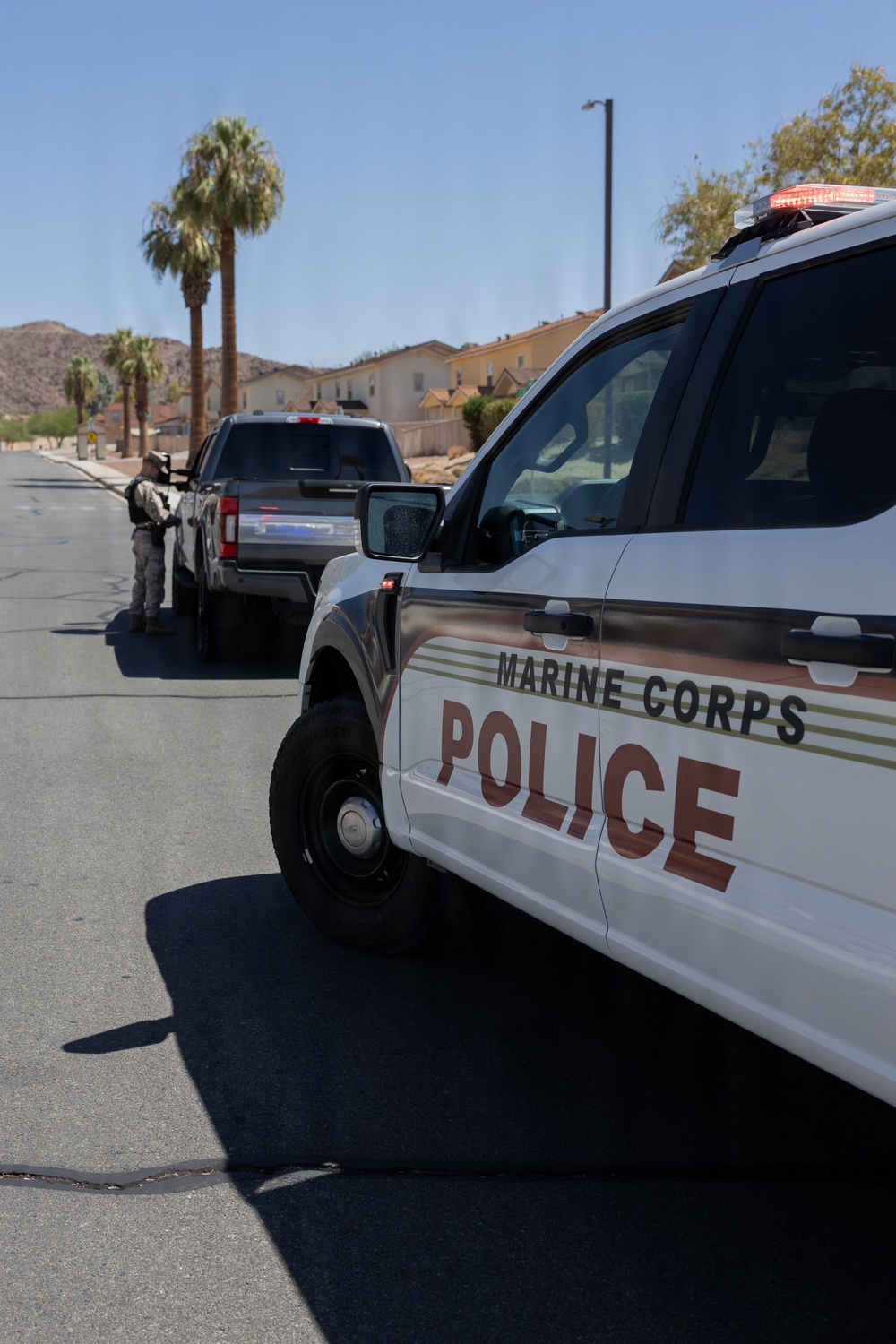 A Marine Corps Police vehicle is prominently displayed in the foreground on a paved road within what appears to be a military base installation. In the background, a uniformed officer is conducting a traffic stop or checkpoint interaction with a dark-colored pickup truck. The setting includes residential-style buildings, palm trees, and rocky hills consistent with a desert military base environment.