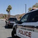 A Marine Corps Police vehicle is prominently displayed in the foreground on a paved road within what appears to be a military base installation. In the background, a uniformed officer is conducting a traffic stop or checkpoint interaction with a dark-colored pickup truck. The setting includes residential-style buildings, palm trees, and rocky hills consistent with a desert military base environment.