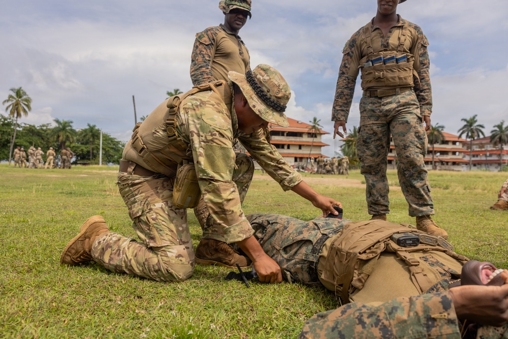 Military personnel are conducting what appears to be a tactical combat casualty care (TCCC) or first aid training exercise on a grass field. One soldier in MultiCam/OCP pattern uniform and boonie hat is kneeling over a simulated casualty lying on the ground, while two others in MARPAT (Marine) uniforms observe. The setting appears to be a tropical or subtropical military installation based on the palm trees and architecture visible in the background.