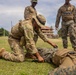 Military personnel are conducting what appears to be a tactical combat casualty care (TCCC) or first aid training exercise on a grass field. One soldier in MultiCam/OCP pattern uniform and boonie hat is kneeling over a simulated casualty lying on the ground, while two others in MARPAT (Marine) uniforms observe. The setting appears to be a tropical or subtropical military installation based on the palm trees and architecture visible in the background.