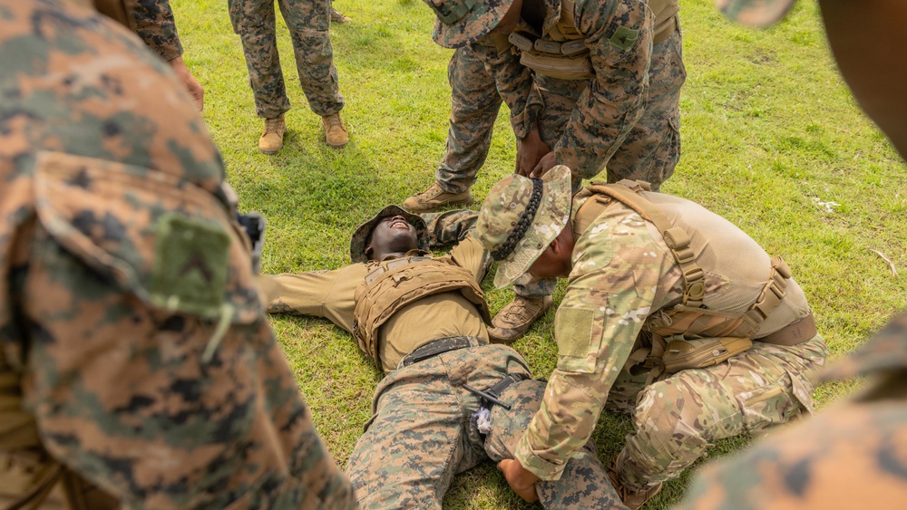 Military personnel in camouflage uniforms are conducting what appears to be a combat casualty care or tactical combat casualty care (TCCC) training exercise outdoors on a grass field. One service member is lying on the ground simulating a casualty while others kneel beside him applying what appears to be a tourniquet or medical aid to his leg. Observers in U.S. Marine Corps MARPAT uniforms stand around the training scenario.