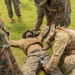 Military personnel in camouflage uniforms are conducting what appears to be a combat casualty care or tactical combat casualty care (TCCC) training exercise outdoors on a grass field. One service member is lying on the ground simulating a casualty while others kneel beside him applying what appears to be a tourniquet or medical aid to his leg. Observers in U.S. Marine Corps MARPAT uniforms stand around the training scenario.