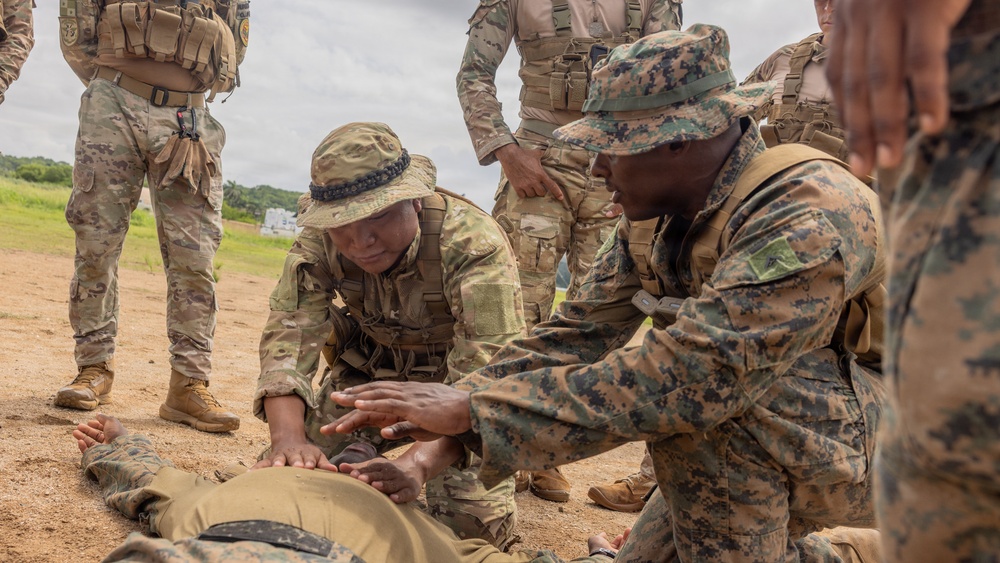 Military personnel in mixed camouflage uniforms are conducting a combat casualty care or tactical first aid training exercise outdoors on a dirt surface. Two soldiers are kneeling over a simulated casualty lying on the ground, with one appearing to instruct the other on medical assessment or treatment techniques. Several other uniformed personnel stand observing in the background.