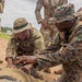 Military personnel in mixed camouflage uniforms are conducting a combat casualty care or tactical first aid training exercise outdoors on a dirt surface. Two soldiers are kneeling over a simulated casualty lying on the ground, with one appearing to instruct the other on medical assessment or treatment techniques. Several other uniformed personnel stand observing in the background.