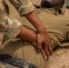 A military service member in OCP (Operational Camouflage Pattern) uniform is performing chest compressions on another person lying on the ground wearing MARPAT (Marine Pattern) digital camouflage uniform. The scene appears to be a combat lifesaver or first aid training exercise conducted on sandy/gravelly terrain. The interlocked hand position is consistent with standard CPR chest compression technique.