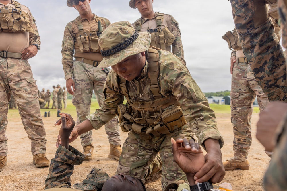 A military training exercise is underway on an open dirt field, where one soldier in MultiCam/OCP uniform and boonie hat is demonstrating or practicing a tactical casualty care or combatives technique on a simulated downed soldier. Several other military personnel in various camouflage uniforms, including MARPAT and OCP patterns, observe the demonstration. The setting appears to be a formal joint training event involving multiple military branches or nations.