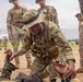 A military training exercise is underway on an open dirt field, where one soldier in MultiCam/OCP uniform and boonie hat is demonstrating or practicing a tactical casualty care or combatives technique on a simulated downed soldier. Several other military personnel in various camouflage uniforms, including MARPAT and OCP patterns, observe the demonstration. The setting appears to be a formal joint training event involving multiple military branches or nations.