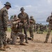 Military personnel in mixed camouflage uniforms are engaged in what appears to be a training exercise on an open field. One soldier is kneeling in a firing or instructional position while others observe, suggesting a demonstration or skills assessment. The tropical environment with palm trees and a red-roofed building in the background indicates a location in a warm, possibly Central or South American region.