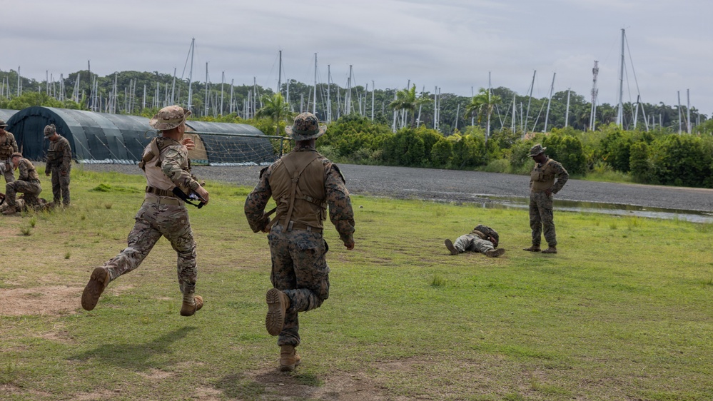 Military personnel in camouflage uniforms are conducting what appears to be a field training exercise on a grassy area. Two soldiers are running toward a prone figure on the ground while another stands nearby observing. The setting includes military structures, netting, and tropical vegetation in the background.