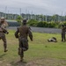 Military personnel in camouflage uniforms are conducting what appears to be a field training exercise on a grassy area. Two soldiers are running toward a prone figure on the ground while another stands nearby observing. The setting includes military structures, netting, and tropical vegetation in the background.