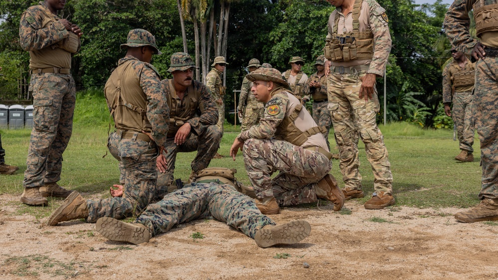 Military personnel in camouflage uniforms are conducting what appears to be a combat casualty care or tactical combat training exercise in an outdoor field setting surrounded by tropical vegetation. One soldier is lying on the ground in a simulated casualty role while two others crouch nearby, apparently practicing medical or tactical assessment procedures. The scene involves multiple soldiers from what appear to be at least two different military units based on their distinct uniform patterns.