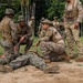 Military personnel in camouflage uniforms are conducting what appears to be a combat casualty care or tactical combat training exercise in an outdoor field setting surrounded by tropical vegetation. One soldier is lying on the ground in a simulated casualty role while two others crouch nearby, apparently practicing medical or tactical assessment procedures. The scene involves multiple soldiers from what appear to be at least two different military units based on their distinct uniform patterns.
