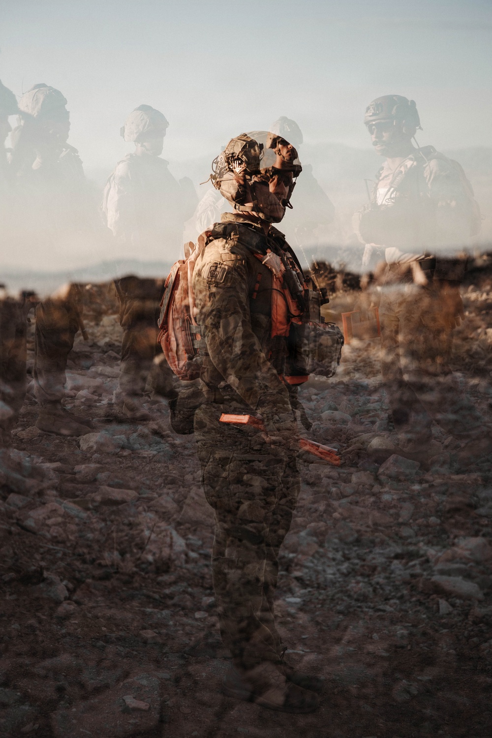A double-exposure or composite photograph depicting a heavily equipped soldier in camouflage gear and tactical helmet standing in a rocky, arid desert landscape. The image layers multiple exposures of soldiers, creating a ghostly, semi-transparent effect with figures visible in the background. The artistic technique conveys themes of multiplicity, memory, or the psychological weight of military service.