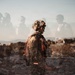 A double-exposure or composite photograph depicting a heavily equipped soldier in camouflage gear and tactical helmet standing in a rocky, arid desert landscape. The image layers multiple exposures of soldiers, creating a ghostly, semi-transparent effect with figures visible in the background. The artistic technique conveys themes of multiplicity, memory, or the psychological weight of military service.