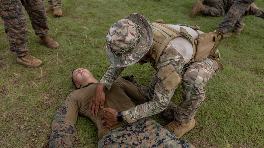 A military training exercise is underway on a grassy field where one service member in OCP/Multicam camouflage, tactical vest, and boonie hat is performing what appears to be a combat casualty care or first aid assessment on another service member in MARPAT (Marine Corps) uniform lying on the ground. The standing/kneeling individual is placing hands on the supine person's torso, consistent with a medical assessment drill. Additional personnel in MARPAT uniforms are visible observing in the background.
