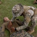 A military training exercise is underway on a grassy field where one service member in OCP/Multicam camouflage, tactical vest, and boonie hat is performing what appears to be a combat casualty care or first aid assessment on another service member in MARPAT (Marine Corps) uniform lying on the ground. The standing/kneeling individual is placing hands on the supine person's torso, consistent with a medical assessment drill. Additional personnel in MARPAT uniforms are visible observing in the background.