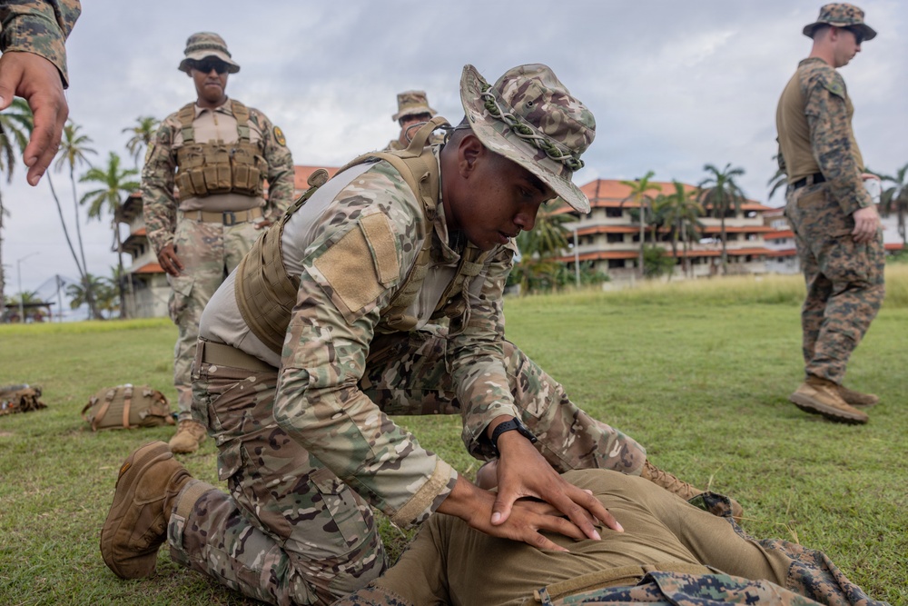 Military personnel are conducting a ground combat or detainee handling training exercise on a grass field. One soldier in MultiCam/OCP camouflage and a boonie hat is kneeling over a prone individual, demonstrating a control or search technique. Several observers in mixed camouflage uniforms, including what appear to be U.S. Marines in MARPAT, watch the demonstration.