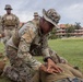 Military personnel are conducting a ground combat or detainee handling training exercise on a grass field. One soldier in MultiCam/OCP camouflage and a boonie hat is kneeling over a prone individual, demonstrating a control or search technique. Several observers in mixed camouflage uniforms, including what appear to be U.S. Marines in MARPAT, watch the demonstration.