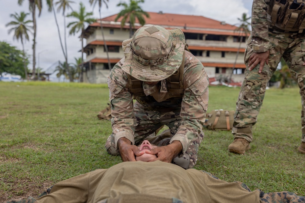 A military service member in MultiCam/OCP camouflage uniform and boonie hat is performing what appears to be a combat casualty care or first aid procedure on a person lying on the ground. The kneeling soldier appears to be conducting airway management or jaw-thrust maneuver on the supine individual. A second uniformed service member stands nearby observing, suggesting this is a training exercise.