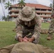 A military service member in MultiCam/OCP camouflage uniform and boonie hat is performing what appears to be a combat casualty care or first aid procedure on a person lying on the ground. The kneeling soldier appears to be conducting airway management or jaw-thrust maneuver on the supine individual. A second uniformed service member stands nearby observing, suggesting this is a training exercise.