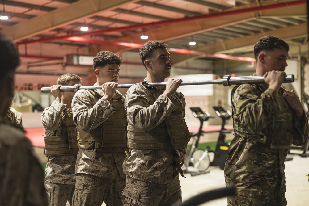 A group of military personnel in camouflage uniforms and tactical vests are conducting a physical training exercise inside what appears to be a military gymnasium or fitness facility. They are performing a barbell carry drill, with multiple individuals sharing the load of a single barbell across their shoulders. The setting includes gym equipment visible in the background, including treadmills and exercise bikes.