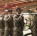A group of military personnel in camouflage uniforms and tactical vests are conducting a physical training exercise inside what appears to be a military gymnasium or fitness facility. They are performing a barbell carry drill, with multiple individuals sharing the load of a single barbell across their shoulders. The setting includes gym equipment visible in the background, including treadmills and exercise bikes.