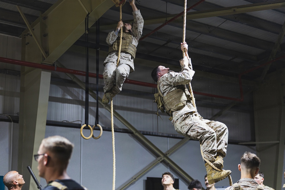 Two military personnel in camouflage uniforms and tactical vests are climbing ropes inside what appears to be a training facility or gymnasium. They are performing a physical fitness exercise while observers watch from below. The setting includes gymnastic rings and an industrial-style building structure.