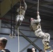 Two military personnel in camouflage uniforms and tactical vests are climbing ropes inside what appears to be a training facility or gymnasium. They are performing a physical fitness exercise while observers watch from below. The setting includes gymnastic rings and an industrial-style building structure.