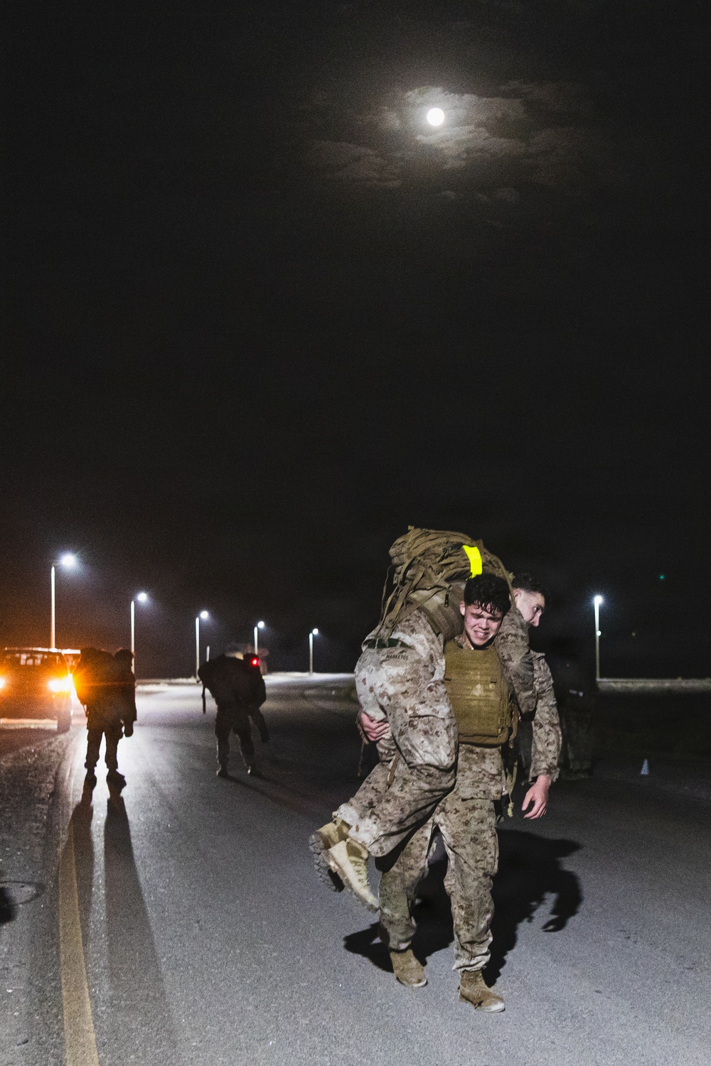 A nighttime military training or operational scene showing personnel in desert camouflage uniforms conducting what appears to be a ruck march or casualty carry exercise on a paved road. The foreground shows one service member carrying another on their back with a large rucksack, illuminated by street lights and a full moon overhead. Additional personnel are visible in the background along the road near vehicles.