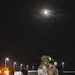A nighttime military training or operational scene showing personnel in desert camouflage uniforms conducting what appears to be a ruck march or casualty carry exercise on a paved road. The foreground shows one service member carrying another on their back with a large rucksack, illuminated by street lights and a full moon overhead. Additional personnel are visible in the background along the road near vehicles.