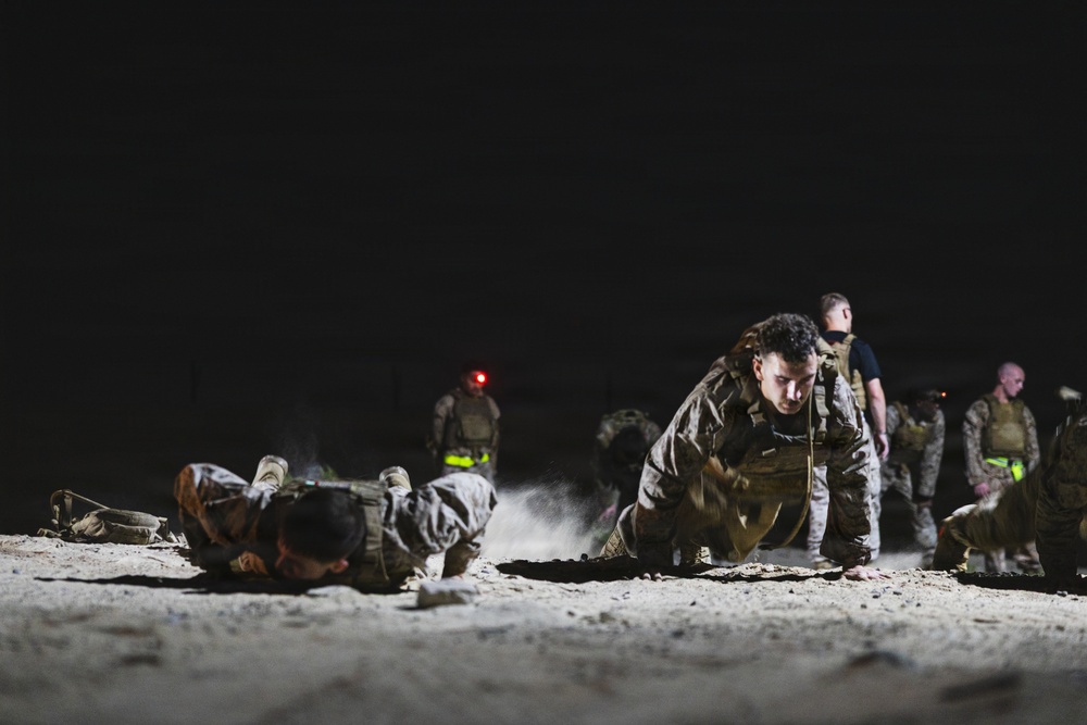 A nighttime military physical training exercise captured under artificial lighting on sandy/dusty terrain. Multiple personnel in camouflage uniforms and tactical vests are performing push-ups or low crawl movements on the ground. At least one individual in a dark shirt and vest appears to be supervising or instructing the group.