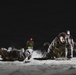 A nighttime military physical training exercise captured under artificial lighting on sandy/dusty terrain. Multiple personnel in camouflage uniforms and tactical vests are performing push-ups or low crawl movements on the ground. At least one individual in a dark shirt and vest appears to be supervising or instructing the group.