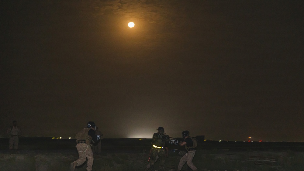 A nighttime scene showing several individuals in military or tactical gear operating in a dark outdoor environment under a bright full moon. The moon illuminates the scene with an orange-amber glow, casting limited light on the figures below. Distant lights are visible on the horizon, suggesting a populated area or base in the background.