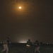 A nighttime scene showing several individuals in military or tactical gear operating in a dark outdoor environment under a bright full moon. The moon illuminates the scene with an orange-amber glow, casting limited light on the figures below. Distant lights are visible on the horizon, suggesting a populated area or base in the background.