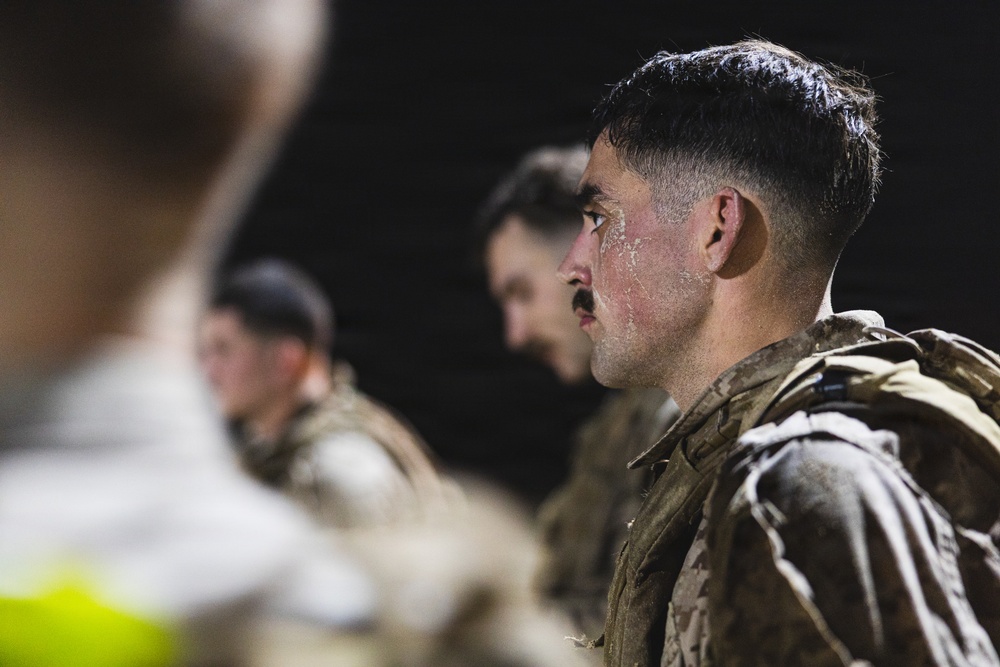 The image shows military personnel in camouflage uniforms standing in a line or formation during what appears to be a nighttime training exercise or event. The foreground subject has visible white residue on his face, suggesting possible exposure to sand, dust, or a training substance. The shallow depth of field keeps the background figures blurred while focusing on the primary subject.