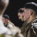 The image shows military personnel in camouflage uniforms standing in a line or formation during what appears to be a nighttime training exercise or event. The foreground subject has visible white residue on his face, suggesting possible exposure to sand, dust, or a training substance. The shallow depth of field keeps the background figures blurred while focusing on the primary subject.