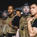 The image shows a group of individuals wearing tactical/plate carrier vests and Bauer hockey helmets with cage face guards, standing in a line at night. They appear to be in a security or crowd-control formation, dressed in black shirts with tan/coyote-colored plate carriers. The setting is dark, suggesting a nighttime outdoor environment.