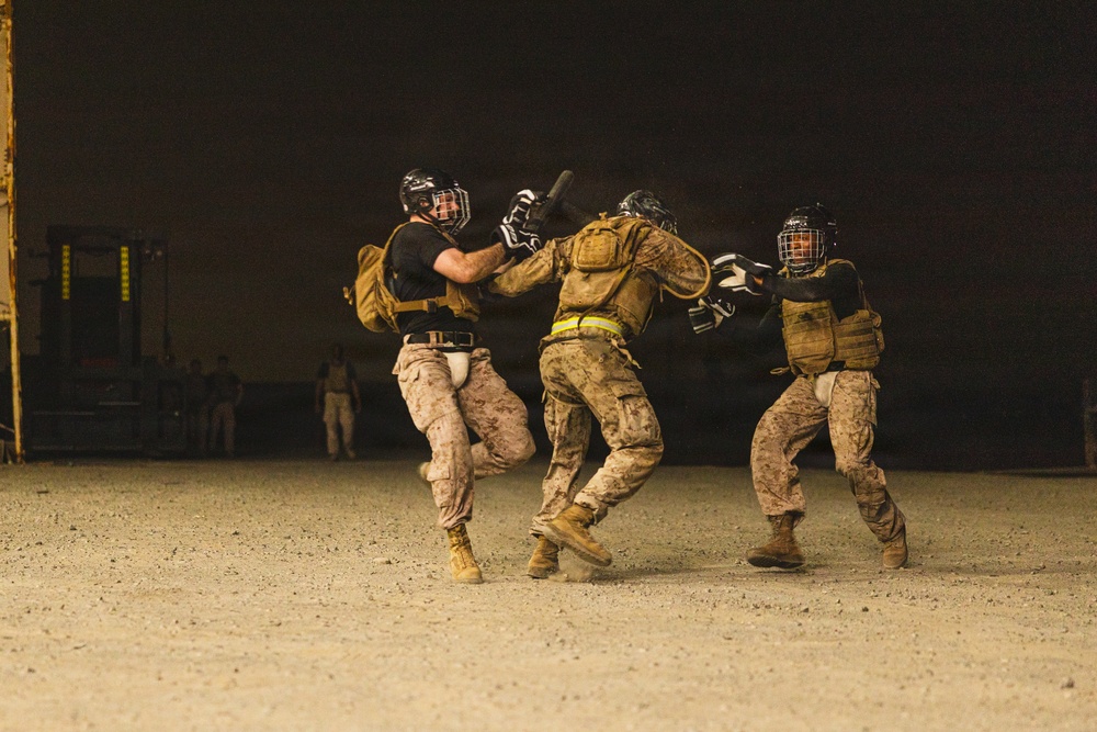 Three individuals in military uniforms and protective gear are engaged in a close-quarters combat training exercise on a sandy, open ground at night. They are wearing helmets with face guards, tactical vests, and padded protection, suggesting a structured martial arts or combatives drill. One participant appears to be wielding a training baton or stick while the others engage in defensive or offensive maneuvers.