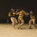 Three individuals in military uniforms and protective gear are engaged in a close-quarters combat training exercise on a sandy, open ground at night. They are wearing helmets with face guards, tactical vests, and padded protection, suggesting a structured martial arts or combatives drill. One participant appears to be wielding a training baton or stick while the others engage in defensive or offensive maneuvers.