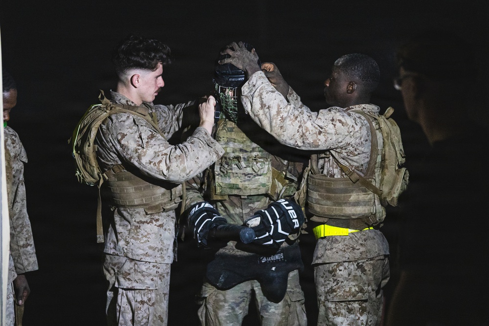 Military personnel in desert camouflage uniforms are assisting a central figure with donning a Bauer hockey helmet/cage at night. Two service members on either side are helping adjust the protective headgear, which appears to be used in a combatives or martial arts training context. Hockey gloves (also Bauer brand) are visible, suggesting a structured hand-to-hand combat or pugil stick training exercise.