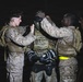 Military personnel in desert camouflage uniforms are assisting a central figure with donning a Bauer hockey helmet/cage at night. Two service members on either side are helping adjust the protective headgear, which appears to be used in a combatives or martial arts training context. Hockey gloves (also Bauer brand) are visible, suggesting a structured hand-to-hand combat or pugil stick training exercise.