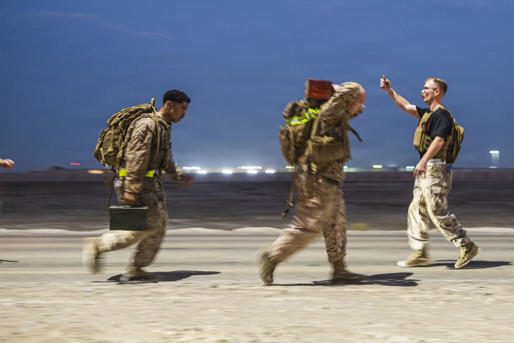 Three military personnel in desert camouflage uniforms are running across what appears to be an airfield or tarmac at night or dusk. Two of the individuals carry large backpacks and equipment including what appears to be an ammunition can, while a third person gestures with a raised hand. The motion blur in the image suggests rapid movement and urgency.