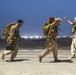Three military personnel in desert camouflage uniforms are running across what appears to be an airfield or tarmac at night or dusk. Two of the individuals carry large backpacks and equipment including what appears to be an ammunition can, while a third person gestures with a raised hand. The motion blur in the image suggests rapid movement and urgency.