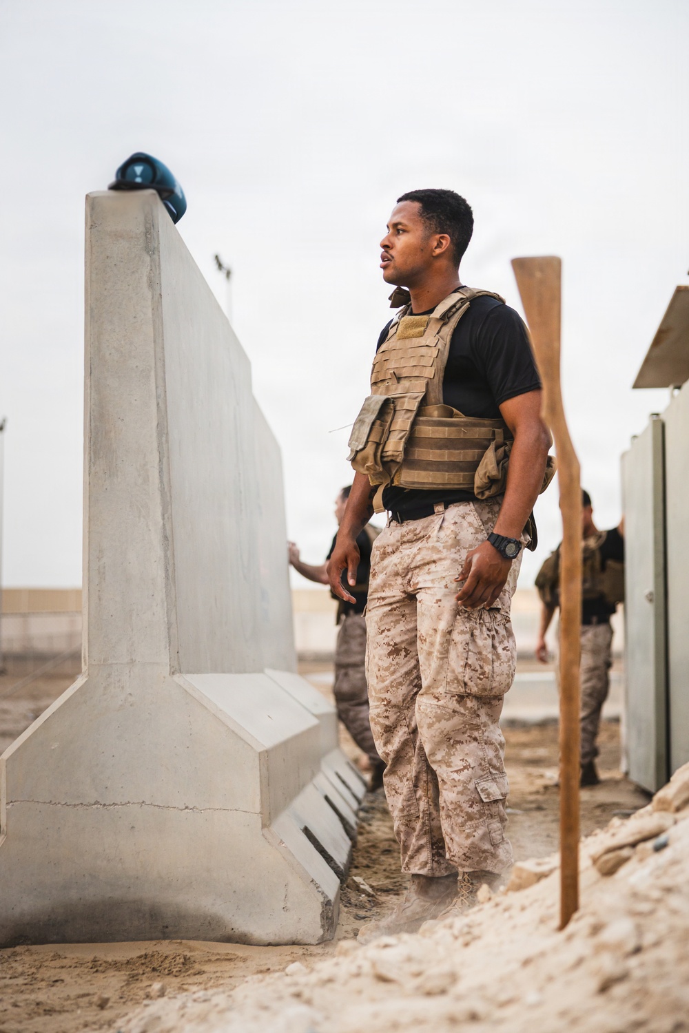 A military or security personnel member stands near a concrete barrier structure in what appears to be an arid, desert environment. He is wearing a tactical plate carrier vest over a black t-shirt with desert camouflage trousers. Additional personnel in similar uniforms are visible in the background.