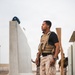 A military or security personnel member stands near a concrete barrier structure in what appears to be an arid, desert environment. He is wearing a tactical plate carrier vest over a black t-shirt with desert camouflage trousers. Additional personnel in similar uniforms are visible in the background.