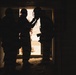 The image shows silhouetted figures standing in a doorway or entrance, backlit by bright exterior light. The figures appear to be wearing tactical or military-style gear including helmets. The strong contrast between the dark interior and bright exterior creates a dramatic silhouette effect.