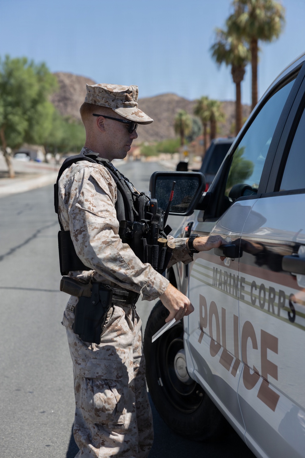 A Marine Corps Police officer in desert MARPAT camouflage uniform is standing beside a Marine Corps Police vehicle, appearing to open or interact with the driver's side door. The officer is equipped with tactical gear including a holstered sidearm, radio, and body armor. The setting appears to be a desert installation environment with palm trees and rocky hills visible in the background.