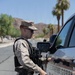 A Marine Corps Police officer in desert MARPAT camouflage uniform is standing beside a Marine Corps Police vehicle, appearing to open or interact with the driver's side door. The officer is equipped with tactical gear including a holstered sidearm, radio, and body armor. The setting appears to be a desert installation environment with palm trees and rocky hills visible in the background.